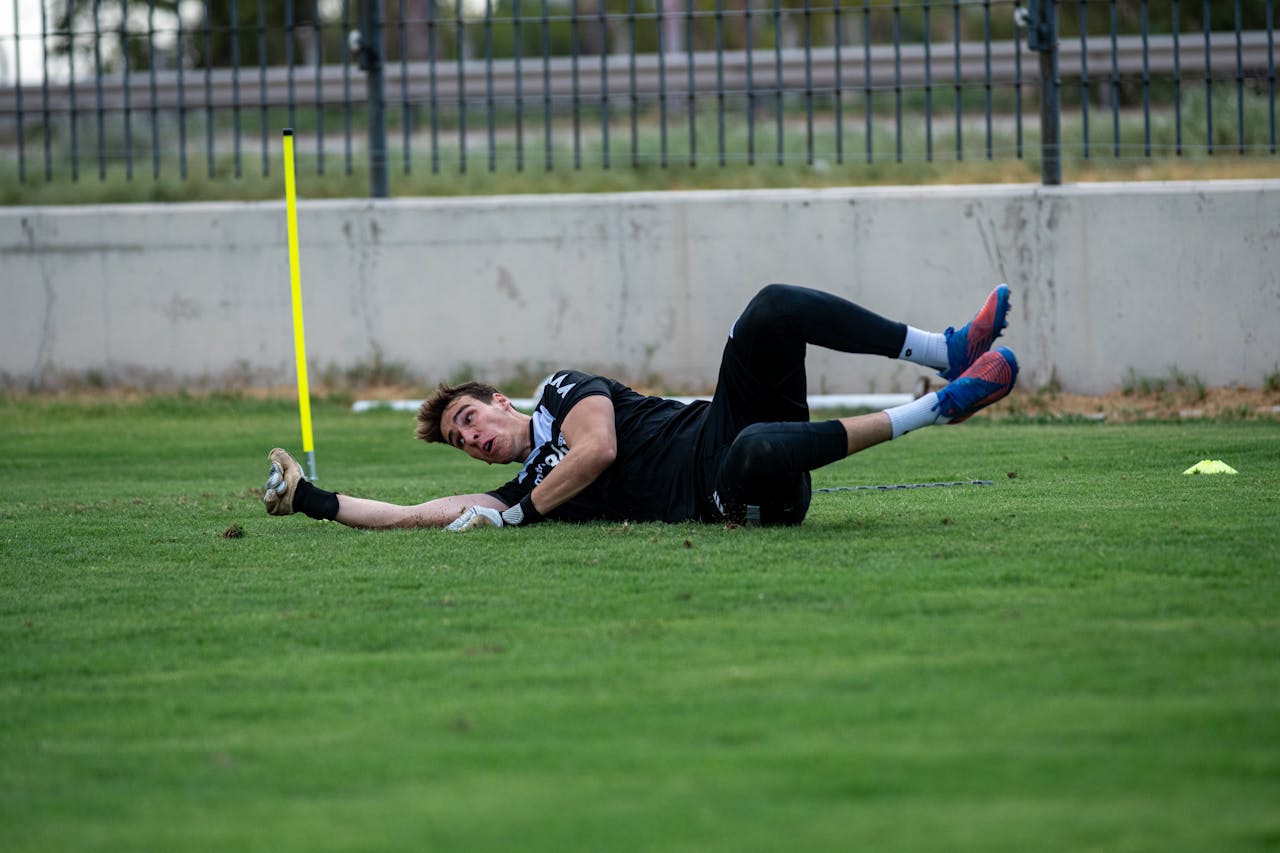 A male goalkeeper in training dives to save a goal on a grassy soccer field.